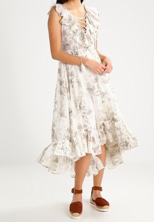 Vrouw draagt een witte midi-jurk met bloemenprint en ruches, een vetersluiting bij de hals en bruine espadrilles met enkelbandje, staand tegen een witte achtergrond.