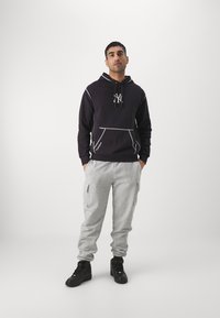 Sudadera oscura con costuras y emblema en gris claro, combinada con pantalones joggers grises claros, que tienen cremalleras laterales y zapatillas negras.