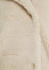 Chaqueta de forro polar color crema con una superficie texturizada, grandes solapas y tres botones color crema en la parte delantera.