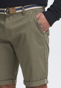 Shorts kaki avec des revers roulés, fabriqués en tissu lisse. Associés à une ceinture rayée beige et bleue, dotée d'une boucle en métal ronde.