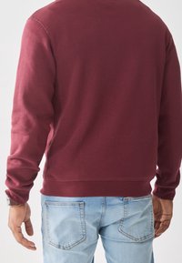 Sudadera de forro polar color granate con cuello redondo y mangas largas, combinada con vaqueros azul claro que presentan bolsillos traseros distintivos.