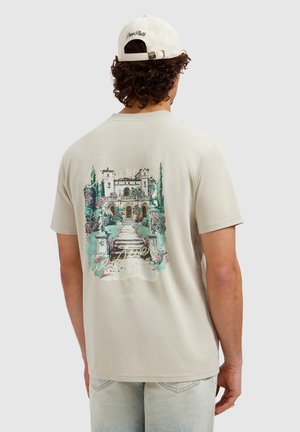 Man draagt beige t-shirt en pet, met de rug naar de camera, t-shirt toont een kleurrijke illustratie van een tuin met trappen en een gebouw.