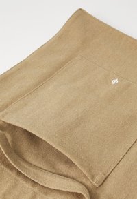 Beige Stoff mit glatter Textur, ausgestattet mit einer rechteckigen Tasche und einem kleinen weißen Logo. Das Design ist einfach und funktional.