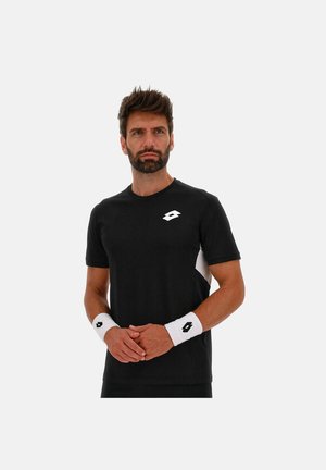 Mann in schwarzem Sport-T-Shirt und weißen Armbändern mit Logos, der vor schlichtem hellem Hintergrund mit verschränkten Händen steht.