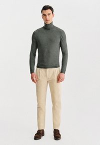 Szary sweter z golfem wykonany z miękkiego dzianiny, zestawiony z beżowymi spodniami i brązowymi butami. Model stoi wyprostowany na jednolitym tle.