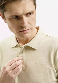 Beżowa koszulka polo z kołnierzykiem, teksturowany materiał i haftowane logo na piersi. Ręka modela poprawia guzik kołnierza.