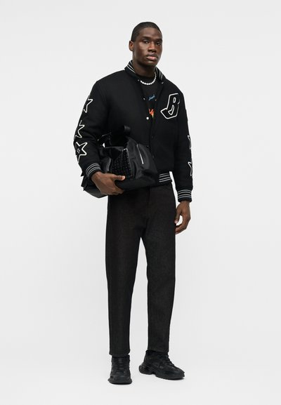 Άνδρας που φοράει μαύρο varsity μπουφάν με αστέρια ραμμένα, μαύρο τζιν, μαύρα αθλητικά παπούτσια και κρατά μια μαύρη τσάντα με καρφιά σε φόντο απλό.