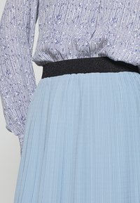Donna che indossa una blusa a maniche lunghe con motivo blu infilata in una gonna plissettata azzurra chiara con una cintura nera.