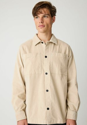 Jonge man draagt een beige corduroy overhemd met knopen en twee borstzakken, kijkend naar de zijkant tegen een effen achtergrond.