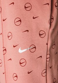 Růžové látkové šortky s opakujícím se vzorem červených kruhových log a Nike swooshů. Design je měkký a ležérní.