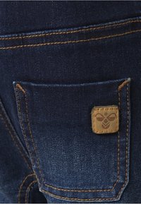 I jeans in denim presentano un tessuto blu scuro con cuciture arancioni a contrasto. Una distintiva toppa quadrata con un design a forma di ape è presente sulla tasca posteriore.