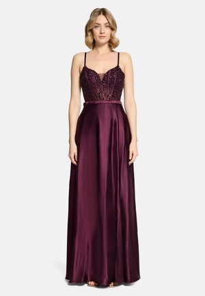 Vrouw draagt een donkergelijke, mouwloze satijnen jurk tot op de vloer met een met kralen versierde lijfje en dunne bandjes, staand tegen een witte achtergrond.