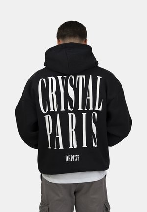 Homme portant un sweat à capuche noir avec un grand texte blanc "CRYSTAL PARIS" et un texte plus petit "DEPT.75" au dos, pantalon gris, tourné de dos.