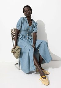 Robe maxi en denim avec des manches ballons, des boutons à l'avant et une taille cintrée. Assortie à des ballerines tressées jaunes et tenant un sac vert.