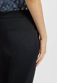 Svarta skräddarsydda byxor, slät tyg, dragkedja i sidofickan, figursydd design, medelhög midja, visad från en sidovy med en hand på midjan.