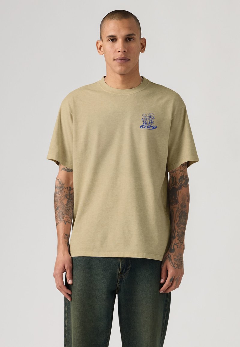 Mann mit Tattoos, der ein beiges Levi’s T-Shirt mit kleinem blauem Cartoon-Motiv auf der Brust und ausgewaschene dunkelgrüne Jeans trägt, vor einem einfarbigen Hintergrund stehend.