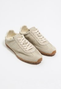 Beige suède sneakers met een getextureerde afwerking, ronde neus en rubberen buitenzool. Heeft veters en een gestikte randdetail op de kuip.