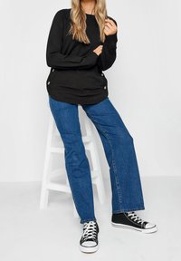 Svart långärmad tröja med sidoknappar, ihop med blå raka jeans och svarta hög-top sneakers. Slätt tyg, avslappnad passform.