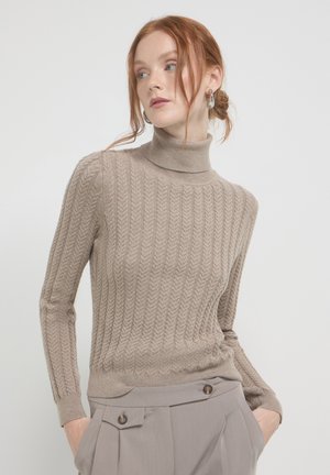 Beige gestrickter Pullover mit hohem Kragen, der ein strukturiertes Fischgrätenmuster und einen gerippten Saum aufweist, kombiniert mit hellgrauen maßgeschneiderten Hosen.