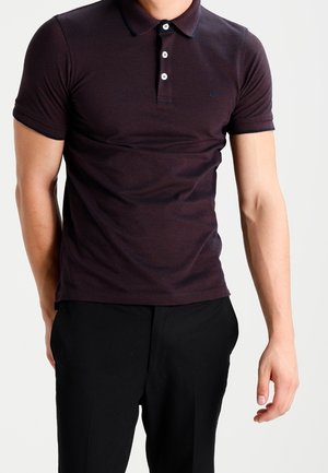 Polo shirt violet foncé au design ajusté, à manches courtes et avec patte de boutonnage à trois boutons. Texture subtile, porté avec un pantalon noir.