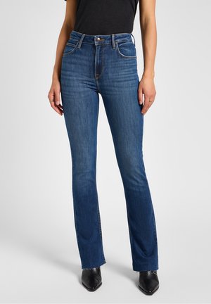 Femme portant un jean bleu foncé ajusté à taille mi-haute avec une légère évasement et un ourlet brut, associé à des bottes noires à bout pointu, debout devant un fond gris.