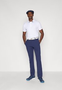 Golfantrekk bestående av en hvit poloskjorte med krage, marineblå bukser og marineblå sneakers med blå detaljer. Modellen har på seg en marineblå caps.