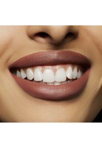 Nærbilde av en smilende munn med hvite tenner og matt mauve leppestift på fyldige lepper. Huden har en nøytral tone og en jevn tekstur.