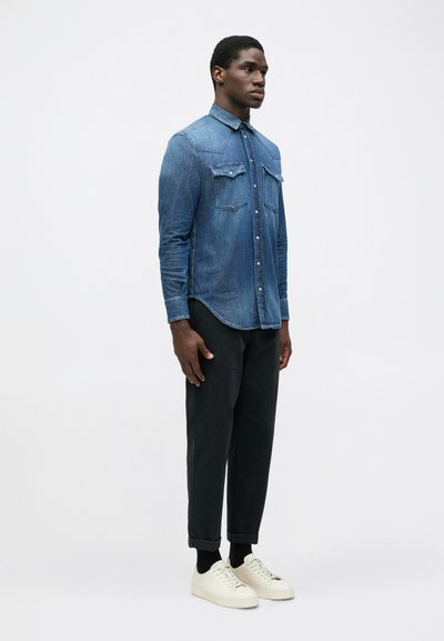 Camicia di denim con colletto a punta, tasche frontali e chiusura con bottoni, abbinata a pantaloni neri tapered e sneakers bianche.