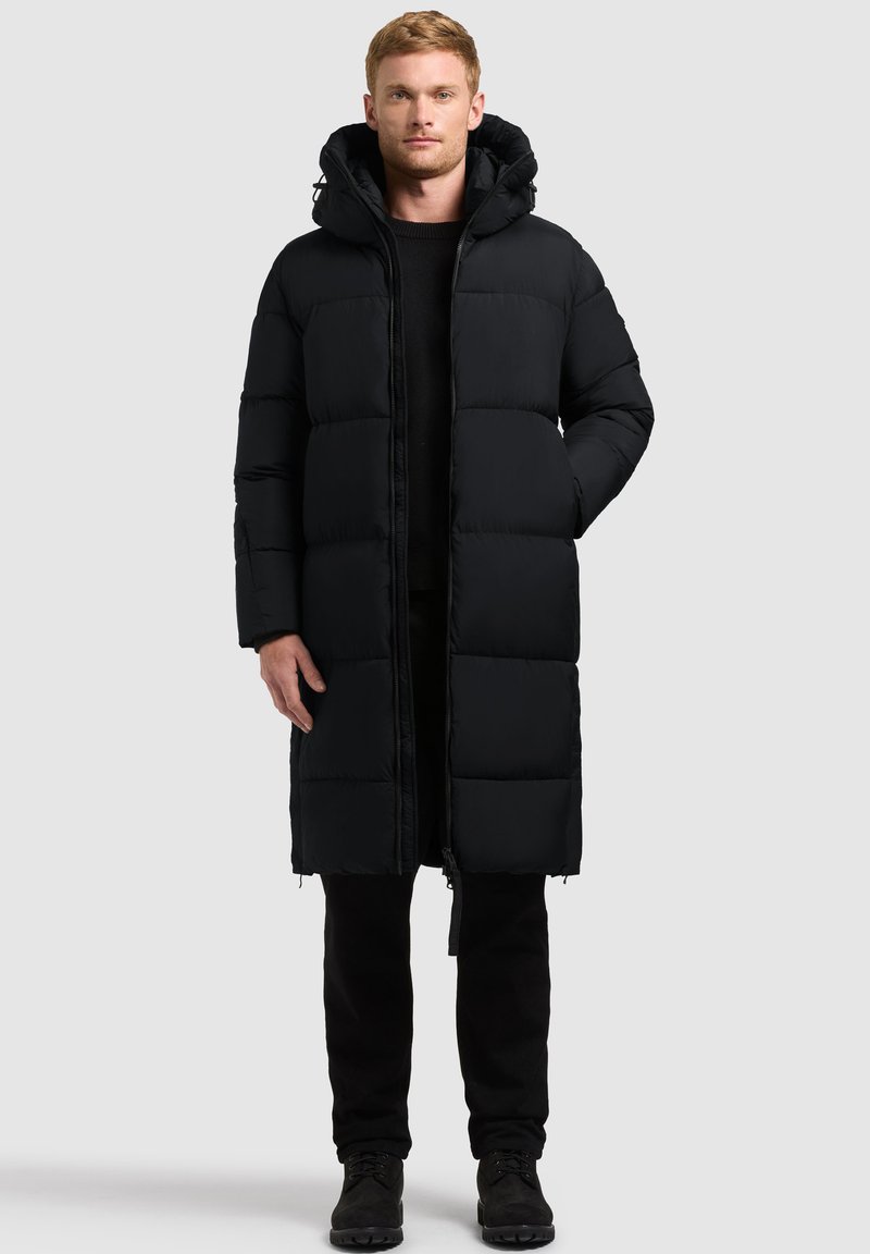 Homme portant un long manteau d'hiver noir matelassé avec une capuche, un pantalon noir et des bottes noires, se tenant face à un fond uni.