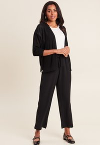 Cardigan nero, con texture e vestibilità ampia, indossato sopra una maglietta bianca. Abbinato a pantaloni a palazzo neri e scarpe basse nere.