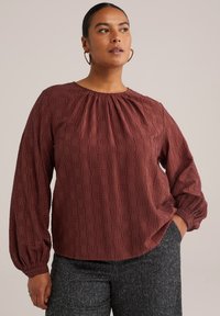 Blusa burdeos con escote fruncido y mangas abullonadas. Patrón texturizado y corte holgado. Combinada con pantalones oscuros texturizados.