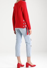 Woman wearing a red sweater with side eyelets and ribbons, light ripped jeans, and red block-heeled shoes on a white background.