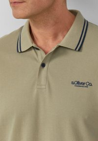 Koszulka polo w jasnooliwkowym kolorze z granatowym, paskowanym kołnierzykiem i granatowym logo. Posiada zapięcie na trzy guziki i teksturowany materiał.