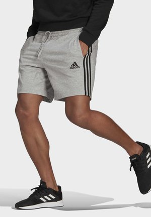 Beine mit grauen Adidas-Shorts mit schwarzen Streifen und schwarzen Adidas-Sneakers, im mittleren Schritt vor einem einfarbigen hellen Hintergrund.