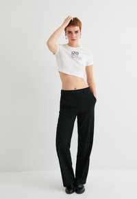 Camiseta corta blanca con estampado gráfico en azul marino y pantalones negros ajustados. El modelo posa con una mano en la cabeza, mostrando una silueta ajustada.
