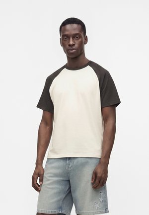 Hombre que lleva una camiseta raglán de manga corta blanca y negra y pantalones cortos vaqueros azul claro con parches de estrellas en la pierna izquierda.