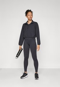 Black zip-up jacket with elastic waist, paired with high-waisted black leggings. Model holds a black water bottle and wears black athletic shoes.