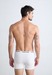 Caleçons-boxer blancs avec une ceinture noire "BJÖRN BORG", design ajusté et texture lisse. Des tatouages visibles couvrent le dos et les bras.