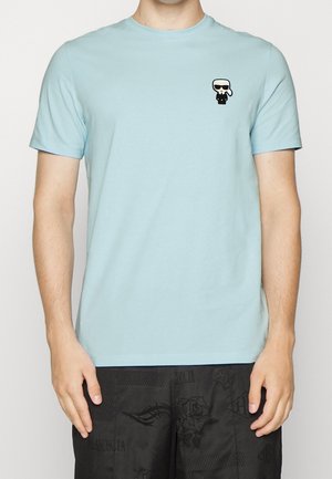 Jasnoniebieska koszulka z krótkim rękawem z małą czarno-białą postacią z kreskówki w okularach przeciwsłonecznych na lewej piersi, noszona z czarnymi szortami w wzory.