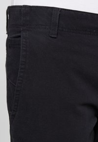 Pantalones de algodón negros con una textura suave y corte ajustado. Presentan bolsillos frontales y detalles de costura sutiles a lo largo de la cinturilla.