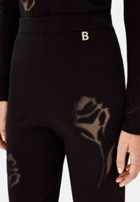 Zwarte broek gemaakt van zachte stof, met doorschijnende bloemenpatronen en een metalen "B"-logo op de tailleband. Versterkte details aan de zijkanten.