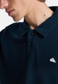 Mørkeblå polo skjorte med et tekstureret mønster, klassisk krave, to-knaps placket og et lille hvidt logo på brystet.