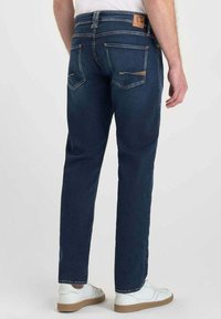 Jeans oscuros de corte recto, con dos bolsillos traseros con costura clara y una etiqueta de cuero en la cintura. Zapatillas blancas visibles.