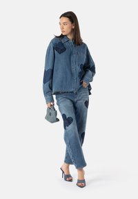 Set di giacca e pantaloni in denim blu con grandi toppe a forma di cuore navy, orli sfilacciati e una borsa blu textured. Le calzature sono mule blu con tacco alto.