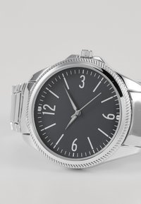 Montre en acier inoxydable avec un cadran noir rond, des index horaires argentés et une lunette texturée. Elle est dotée d'un bracelet en métal et présente un design classique.