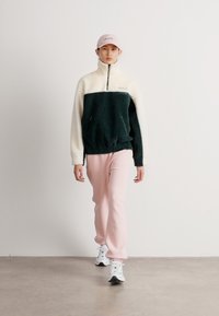 Pulover jakna s flis teksturom u tamnozelenoj i krem boji, s dizajnom na pola zipa. Uparena s svijetloružičastim trenirkama i bijelim tenisicama.