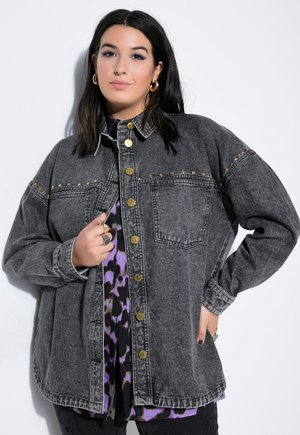 Kvinde med sort hår iført en mørkegrå denimjakke med guldknapper og lilla mønstret skjorte, poserer med hånden på hoften mod hvid baggrund.