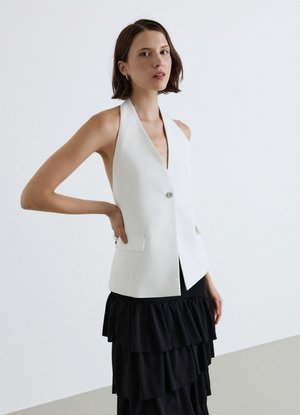 Giovane donna che indossa un gilet bianco sartoriale senza maniche con bottoni e una gonna nera a balze a più livelli, in piedi davanti a uno sfondo chiaro e semplice.