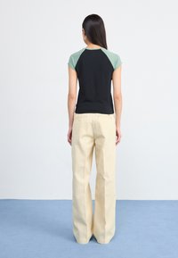 A woman with long dark hair wears a black and green shirt and light beige pants, standing on a light blue rug against a pale background.