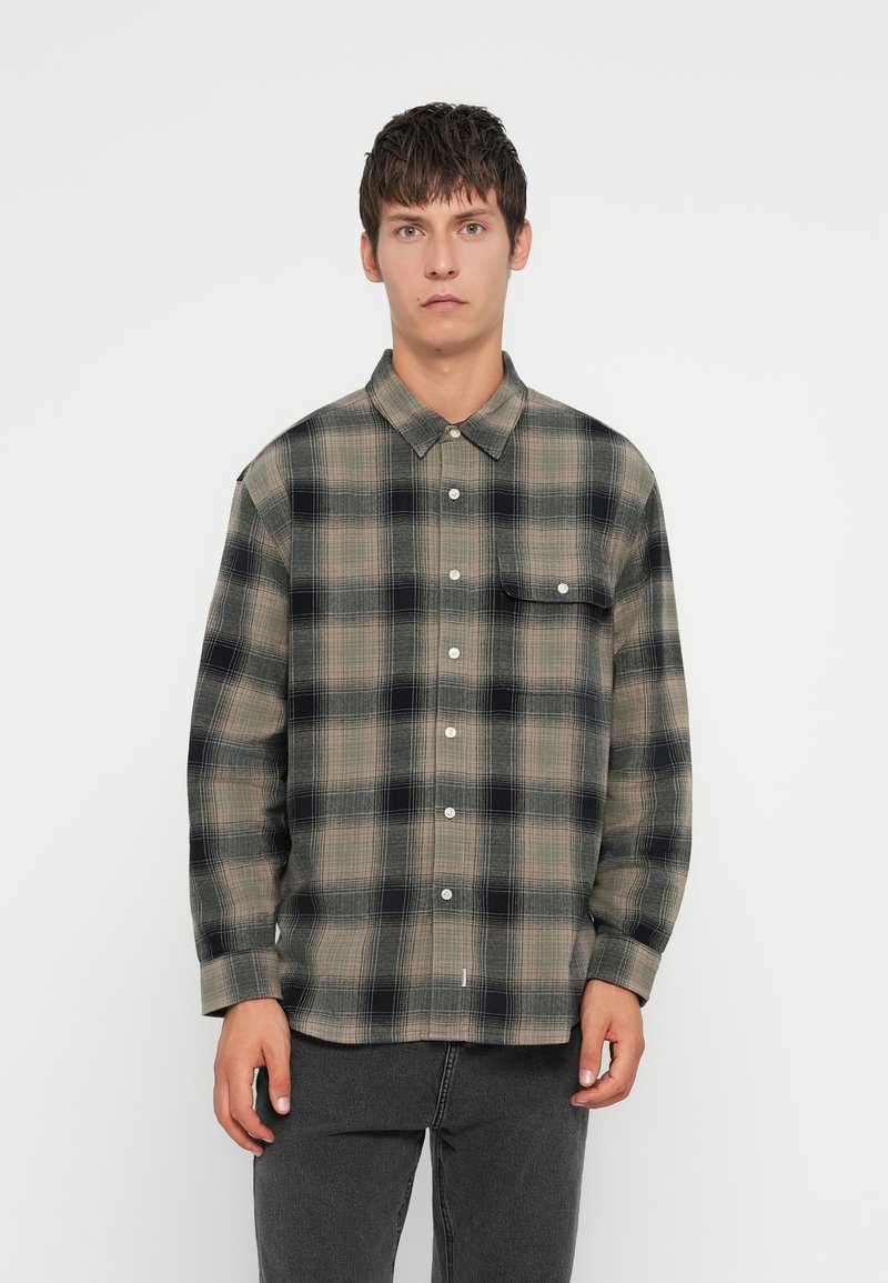 Camisa de franela a cuadros en beige y negro con un diseño de botones, un bolsillo en el pecho y mangas largas. Textura tejida, ajuste holgado.